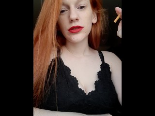 A girl with long red hair in a black dress and red lipstick smokes a cigarette with a brown filter