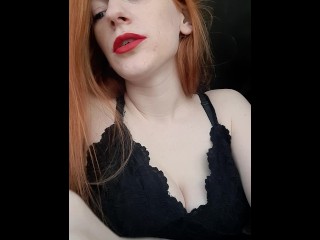 A girl with long red hair in a black dress and red lipstick smokes a cigarette with a brown filter